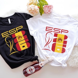May include: Black and white t-shirts with the letters "ESP" and a graphic of the Spanish flag. The shirts are knotted at the waist and are on a white fluffy surface. A bottle of red wine is in the foreground.