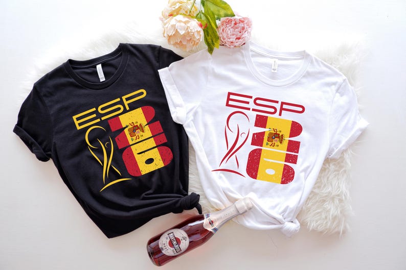 May include: Black and white t-shirts with the letters "ESP" and a graphic of the Spanish flag. The shirts are knotted at the waist and are on a white fluffy surface. A bottle of red wine is in the foreground.