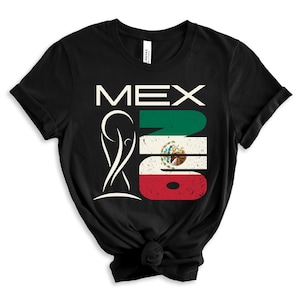 May include: Black t-shirt featuring the word "MEX" in white, with a stylized graphic of a trophy and the Mexican flag. The flag design is incorporated into the numbers "2018". The shirt is made of a soft material.