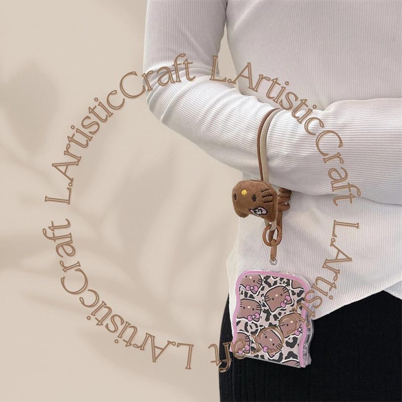 May include: A pink and brown phone case with a Hello Kitty design. The case is attached to a brown and white lanyard with a brown Hello Kitty charm.