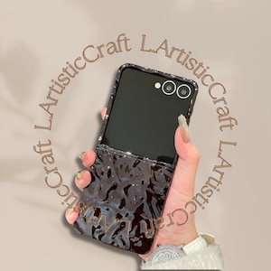 May include: Black phone case with a textured, glossy finish. The case is open, revealing the phone's screen and camera. The case has a glittery border and the text "L.ArtisticCraft" is visible.