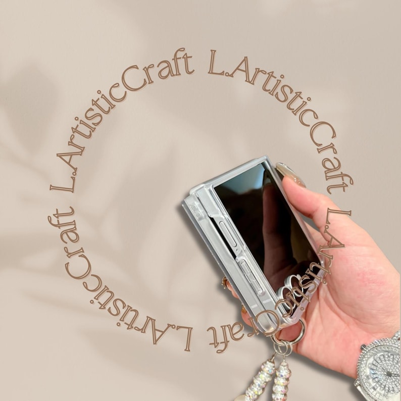 Pu&ograve; includere: Custodia trasparente per telefono con un ciondolo decorativo color argento e un cinturino da polso con perline. La custodia &egrave; tenuta in mano, con un orologio visibile sullo sfondo. Il testo "L.ArtisticCraft" &egrave; in cerchio.