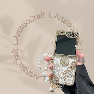 May include: A white and gold phone case with a floral design. The case has a flip-up cover and a pearl chain attached.