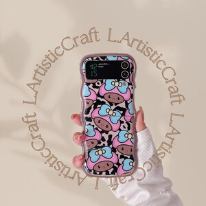 May include: A pink and black phone case with a cartoon character design. The case has a raised edge and a clear back. The phone case is decorated with a pattern of pink and blue bows and brown faces.