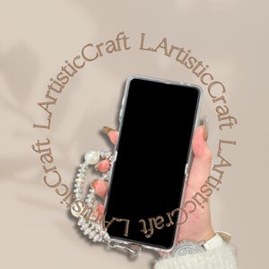Pu&ograve; includere: Una custodia trasparente per telefono con schermo nero &egrave; tenuta in mano. La custodia &egrave; adornata da un cinturino da polso con perle e strass. Lo sfondo &egrave; di colore marrone chiaro con il testo "L.ArtisticCraft" in un motivo circolare.
