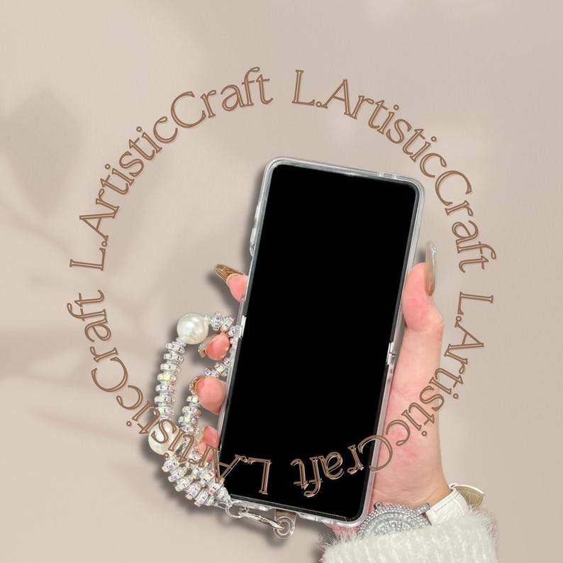 Pu&ograve; includere: Una custodia trasparente per telefono con schermo nero &egrave; tenuta in mano. La custodia &egrave; adornata da un cinturino da polso con perle e strass. Lo sfondo &egrave; di colore marrone chiaro con il testo "L.ArtisticCraft" in un motivo circolare.