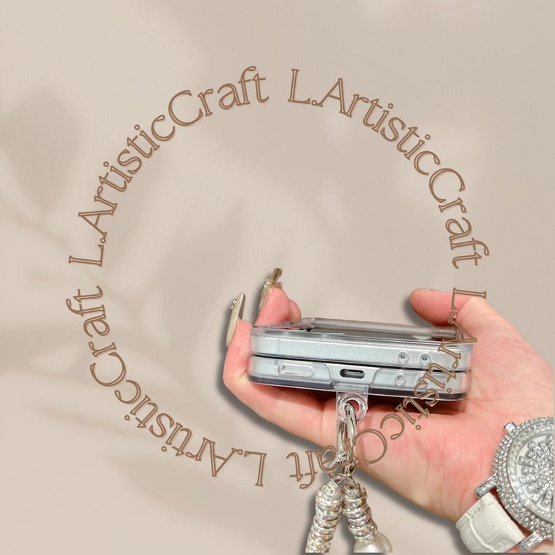 Pu&ograve; includere: Una custodia per telefono argentata con un portachiavi attaccato. La custodia &egrave; tenuta in mano, con il testo "L.ArtisticCraft" in un motivo circolare attorno al telefono. Un ciondolo argentato pende dal portachiavi.
