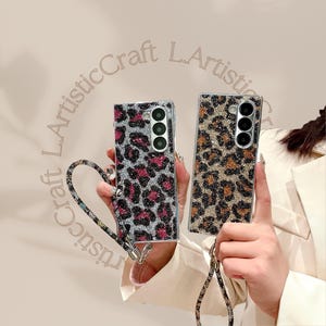 Könnte beinhalten: Zwei verzierte Handyhüllen mit Leopardenmuster. Eine Hülle hat eine silberne Basis mit rosa und schwarzen Flecken, die andere eine goldene Basis mit braunen und schwarzen Flecken. Beide Hüllen haben ein Kameramodul und einen passenden Riemen.