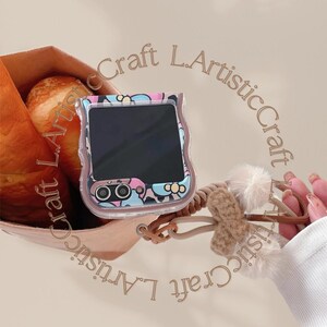 May include: A clear phone case with a colorful floral pattern. The case has a brown strap with a fluffy white pom-pom attached. The text "Craft L.Artistic Craft L.Artistic" is written around the case.