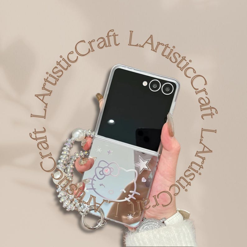 Pu&ograve; includere: Una custodia trasparente per telefono con un design di Hello Kitty, stelle e la scritta "Artistic Craft". La custodia &egrave; attaccata a un cinturino da polso con perline, perle e dettagli rosa. Il telefono &egrave; aperto, mostrando uno schermo nero.