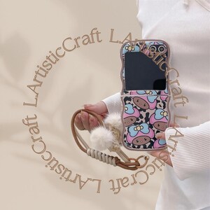 May include: A pink and brown phone case with a cartoon character design. The case has a mirror on the front and a strap with a fluffy pom-pom.