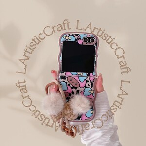 May include: A pink, blue, and brown patterned phone case with a flip-up screen. The case has a cartoon character design with a brown bear face and a pink bow. The phone case has a brown strap with a fluffy white pom-pom attached.