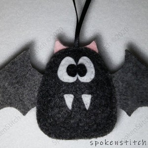 Peut inclure: Une décoration de chauve-souris en feutre noir avec des oreilles roses, des yeux blancs et des crocs blancs. La chauve-souris a une boucle de ruban noir pour l'accrocher.