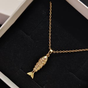 May include: A gold chain necklace with a small gold fish pendant. The fish has a red gemstone for an eye.