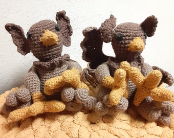 May include: Two brown crocheted amigurumi figures with yellow beaks and feet sitting on a textured tan surface. The figures have large ears and are sitting with their legs crossed.