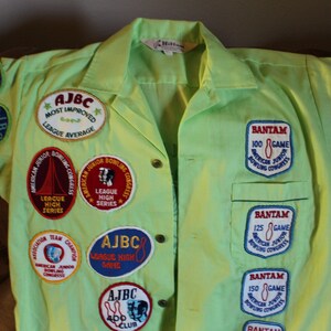 May include: A lime green bowling shirt with multiple embroidered patches. The patches feature bowling pins, bowling balls, and text such as "AJBC", "League High Series", and "Bantam 100 Game".