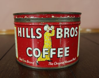 May include: Vintage Hills Bros Coffee tin, red with white lettering and a cartoon figure. The tin reads "Red Can Brand" and "The Original Vacuum Pack". The coffee tin is on a dark wooden surface.