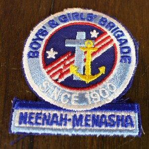 May include: A blue and white embroidered patch with the text "Boys & Girls' Brigade Since 1960" and "Neenah-Menasha". The patch features a red, white, and blue American flag, a yellow anchor, and a white cross.