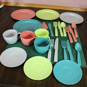 May include: A set of colorful plastic toy dishes and cutlery, including plates, cups, saucers, forks, knives, and spoons. The set is arranged on a green placemat.