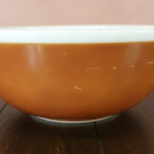 May include: A vintage mixing bowl with a warm, orange-brown exterior and a white rim. The bowl has a rounded shape and appears to be made of ceramic or a similar material. It is likely used for food preparation and baking.