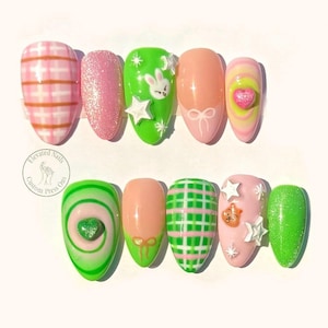 May include: A set of ten press-on nails featuring diverse designs including pink and white plaid, glittery pink, bright green with a white rabbit and stars, peach with a white bow, and pink swirls with a heart. Includes a circular logo.