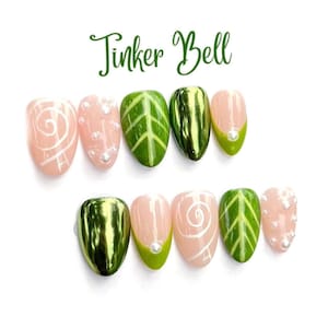 May include: A set of ten press-on nails with the text Tinker Bell at the top. The designs include light pink nails with white swirls or small white pearls, green nails with leaf vein patterns, and solid metallic green nails with a glossy finish.