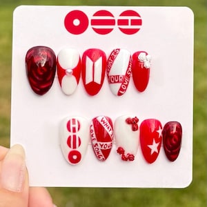May include: A set of ten press-on nails with a variety of designs. The nails feature red, white, and clear polish with accents like a pearl, text that reads "WHAT IS YOUR LOVE SONG?", and a star. The nails are held on a white card.