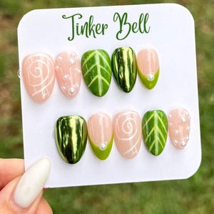 May include: A set of ten press-on nails on a white display card labelled Tinker Bell. The designs include nude nails with white swirls or small pearls, green nails with leaf vein patterns, and solid metallic green nails with a glossy finish.