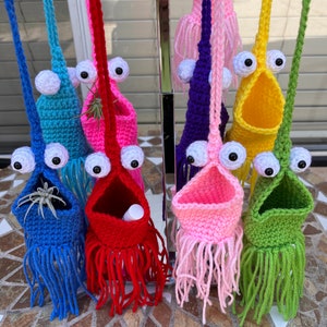 May include: Seven crocheted hanging planters with googly eyes and fringe. The planters are in various colours, including blue, red, pink, yellow, and green. Each planter has a small opening at the top for plants or other small items.