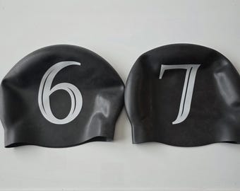 May include: Two black swim caps with white numerals. One cap displays the number 6, and the other displays the number 7. The caps are made of a smooth, flexible material, likely silicone or rubber, and are designed to fit snugly over the head.