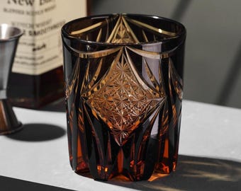 May include: A brown and amber-colored, patterned rocks glass. The glass has a diamond-shaped design in the center and is sitting on a white surface. A bottle and a silver jigger are in the background.