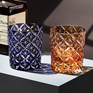 Puede incluir: Dos vasos de whisky de cristal, uno azul oscuro y otro ámbar, con un patrón tallado en diamante. Los vasos están sobre una superficie blanca, con una botella de whisky en el fondo.