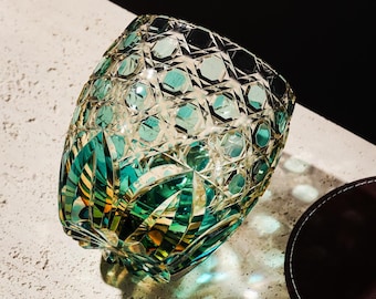 Könnte beinhalten: Ein geschliffener Glasbecher mit geometrischem Muster. Das Glas hat klare und türkis farbene Facetten, die ein Spiel von Licht und Schatten erzeugen. Der Becher steht auf einer strukturierten Oberfläche, mit einem dunkelbraunen Untersetzer.