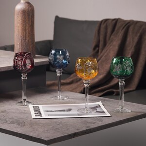 May include: Four coloured crystal wine glasses with starburst and leaf patterns. The glasses are in red, blue, yellow, and green, each with a clear stem. A magazine with the text "JIL SANDER" is on the table.