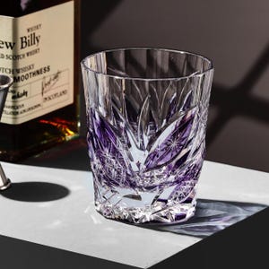 Puede incluir: Un vaso de whisky de cristal tallado con un diseño floral púrpura. El vaso es cilíndrico con una base gruesa y está parcialmente lleno de un líquido oscuro. El vaso está sobre una superficie blanca.