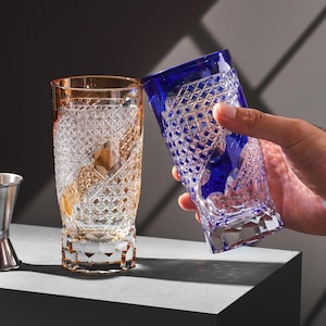 Puede incluir: Dos vasos altos estampados. Uno es de color ámbar, el otro es azul. Ambos vasos tienen un diseño de corte de diamante. Una mano sostiene el vaso azul. Los vasos están sobre una superficie gris.
