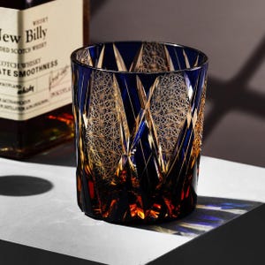 May include: A blue and gold-coloured whiskey glass with a geometric pattern. The glass has a thick base and a wide mouth. The glass is sitting on a white surface. The background includes a bottle of whiskey.