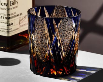 Könnte beinhalten: Ein blau-goldenes Whiskyglas mit geometrischem Muster. Das Glas hat einen dicken Boden und eine weite Öffnung. Das Glas steht auf einer weißen Oberfläche. Der Hintergrund zeigt eine Whiskyflasche.