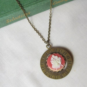 May include: A bronze necklace with a cameo pendant. The cameo features a white silhouette of a couple against a red background. The pendant is surrounded by a decorative bronze circle.