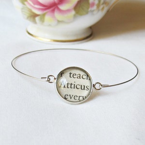 May include: A silver bangle bracelet with a round glass charm featuring the text "r teach Atticus every".
