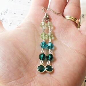 May include: Dangle earrings with a colour gradient of green and teal beads. The earrings have faceted beads in light green, teal, and dark green, connected by silver-coloured metal chains. Each earring's base has two dark green bezel-set stones.