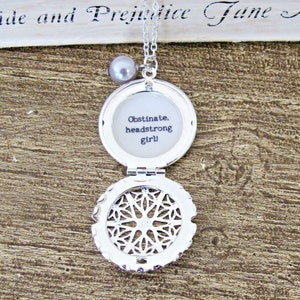 May include: Silver locket necklace with a pearl charm. The locket is open and reveals a white background with the text "Obstinate, headstrong girl!"