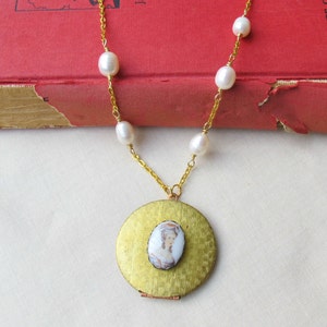 Può includere: Una collana con un ciondolo in oro e un cammeo bianco che raffigura una donna. La collana è realizzata con una catena in oro e dettagli di perle bianche.