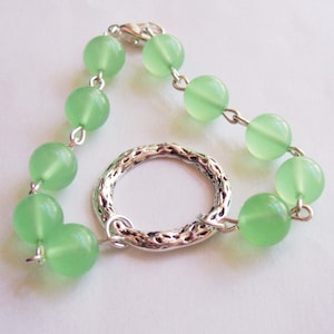 May include: A green glass bead bracelet with a silver charm. The bracelet is made of small, round beads and has a silver ring charm with a textured design.