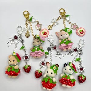 May include: Keychains featuring miniature animal figures dressed in strawberry-themed outfits. Each keychain has a gold or silver clasp, decorative charms, and a sparkling red strawberry charm. The animals include a bear, a cat, a bunny, and a fox.