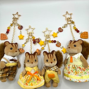 May include: Four flocked squirrel figurines attached to gold-toned star-shaped keychains. Each keychain features dangling charms including yellow stars, autumn-colored beads, maple leaves, and waffle shapes on delicate gold chains.
