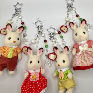 May include: Four flocked rabbit figurines attached to silver star-shaped keychains. Each keychain features a chain with small beads, a red apple charm, and a silver bow. The rabbits wear colorful fabric clothing including dresses and shirts.