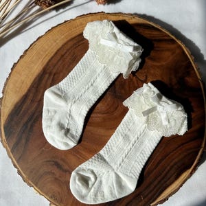 May include: A pair of white baby socks with a cable knit pattern and lace trim. Each sock has a small white bow at the top. The socks are displayed on a round, wooden surface.