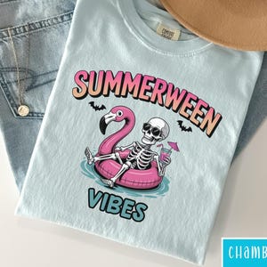May include: Light blue t-shirt with the words "SUMMERWEEN VIBES" and a graphic of a skeleton in sunglasses floating on a pink flamingo pool float. The shirt is folded on top of blue jeans, a necklace, and a tan hat.