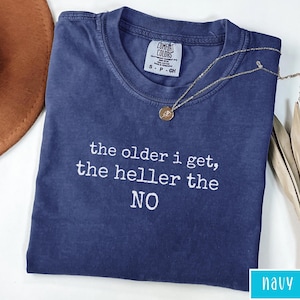 May include: Navy blue t-shirt with the text "the older i get, the heller the NO" in white. A gold necklace with a pendant is draped across the shirt. The shirt has a crew neck and a Comfort Colors tag.
